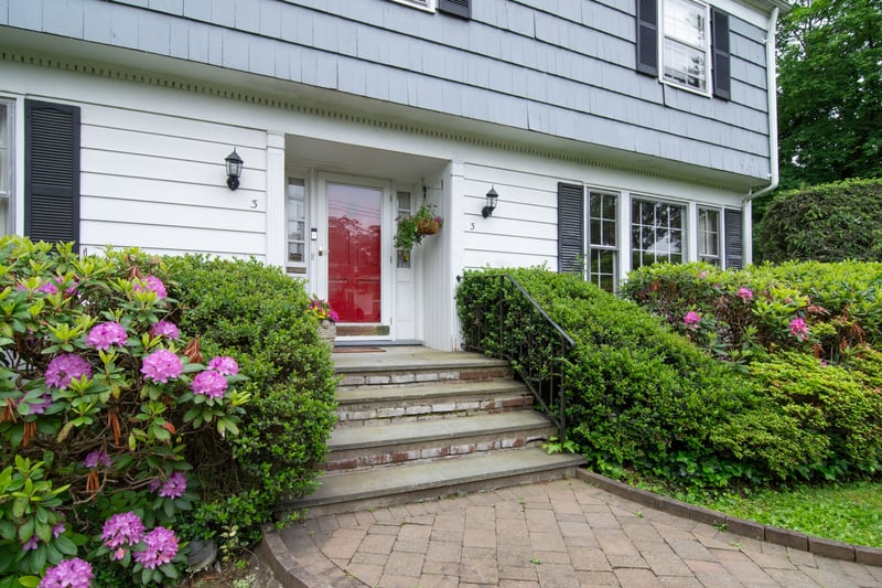 The front entrance is framed by flowering bushes and lush, mature greenery, creating a warm and welcoming first impression.